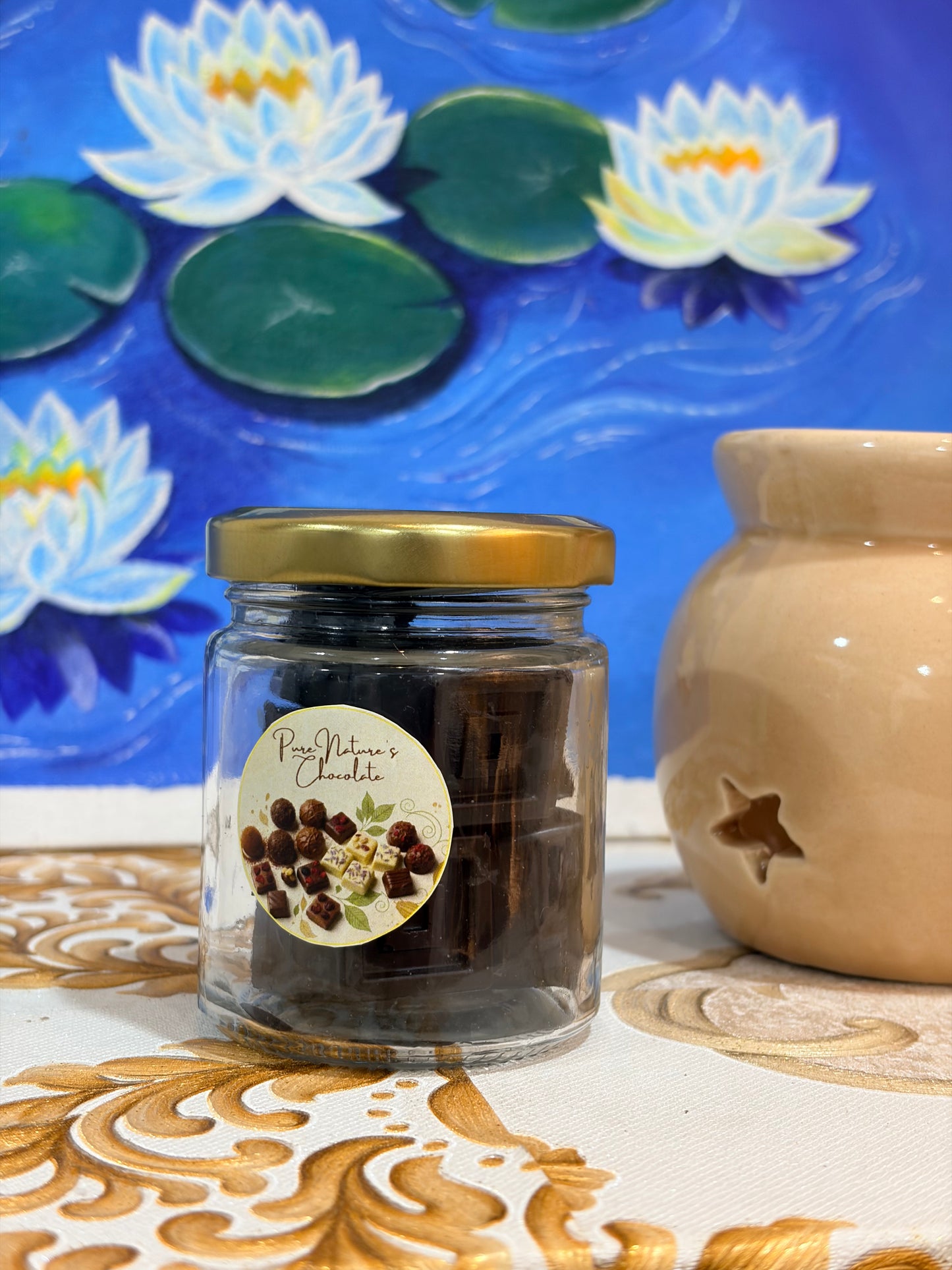 A glass jar of Peony Nature's Chocolate bark with a gold lid and a round peony-themed label, set on a patterned tablecloth next to a ceramic candle holder against a water lily painting background.