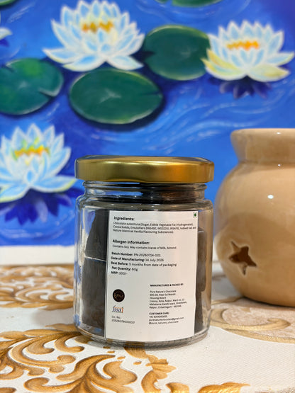 A clear glass jar of dark Chocolate with a gold lid and a white label, placed on a patterned beige surface in front of a blue lotus painting and a beige ceramic pot.